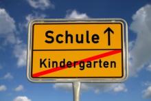 Schild im Stil einer Orsttafel - unten durchgestrichen "Kindergarten", darüber "Schule" mit einem geradeaus zeigenden Pfeil