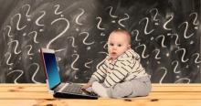 Kleinkind an einem Laptop mit fragendem Blick, im Hintergrund eine Tafel mit vielen Fragezeichen