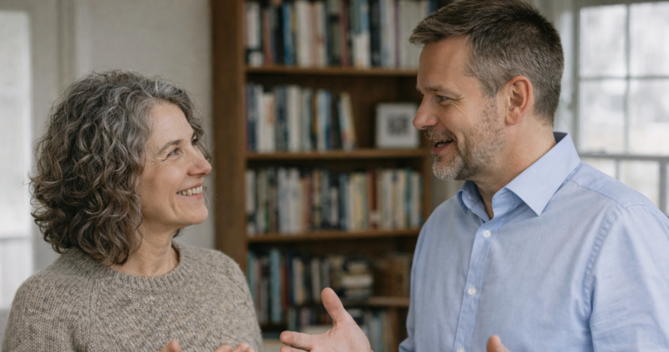 Zwei erwachsene Personen stehen einander in einem hellen Raum gegenüber und führen ein Gespräch. Die Frau mit grauen, lockigen Haaren trägt einen beigen Strickpullover und lächelt den Mann an, während sie eine Hand leicht erhoben hat. Der Mann mit kurzen grauen Haaren und Bart trägt ein hellblaues Hemd und gestikuliert offen mit beiden Händen. Im Hintergrund sind ein Bücherregal und ein Fenster zu sehen. Die Körpersprache beider wirkt aufmerksam und wertschätzend.