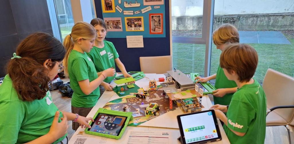 Kinder stehen um einen Tisch, auf dem Tisch befinden sich ihre Modelle aus Klemmbausteinen und Tablets